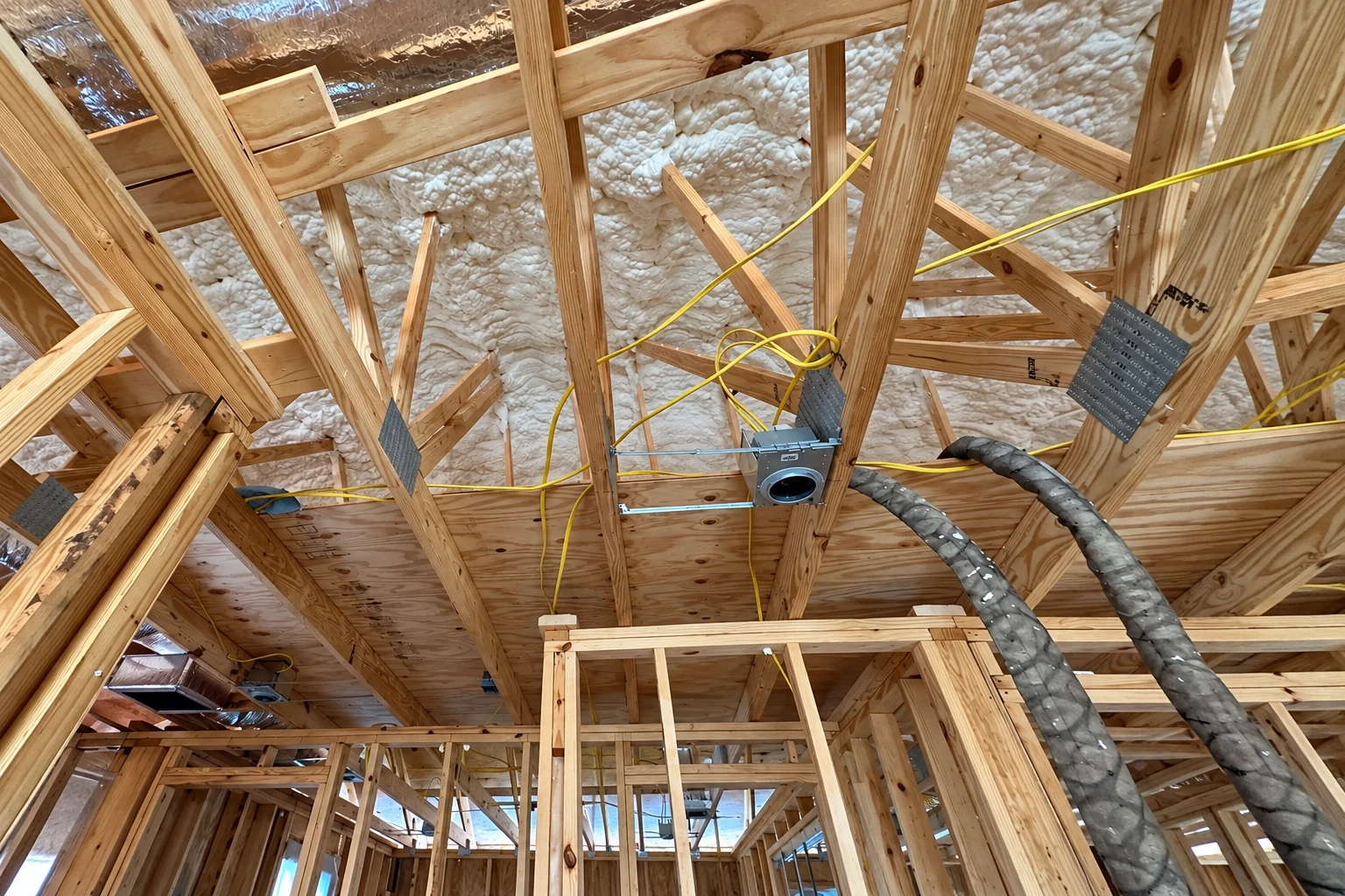 Exposed roof framing with white foam between rafters, yellow wiring, and a spray hose extending toward the ceiling.