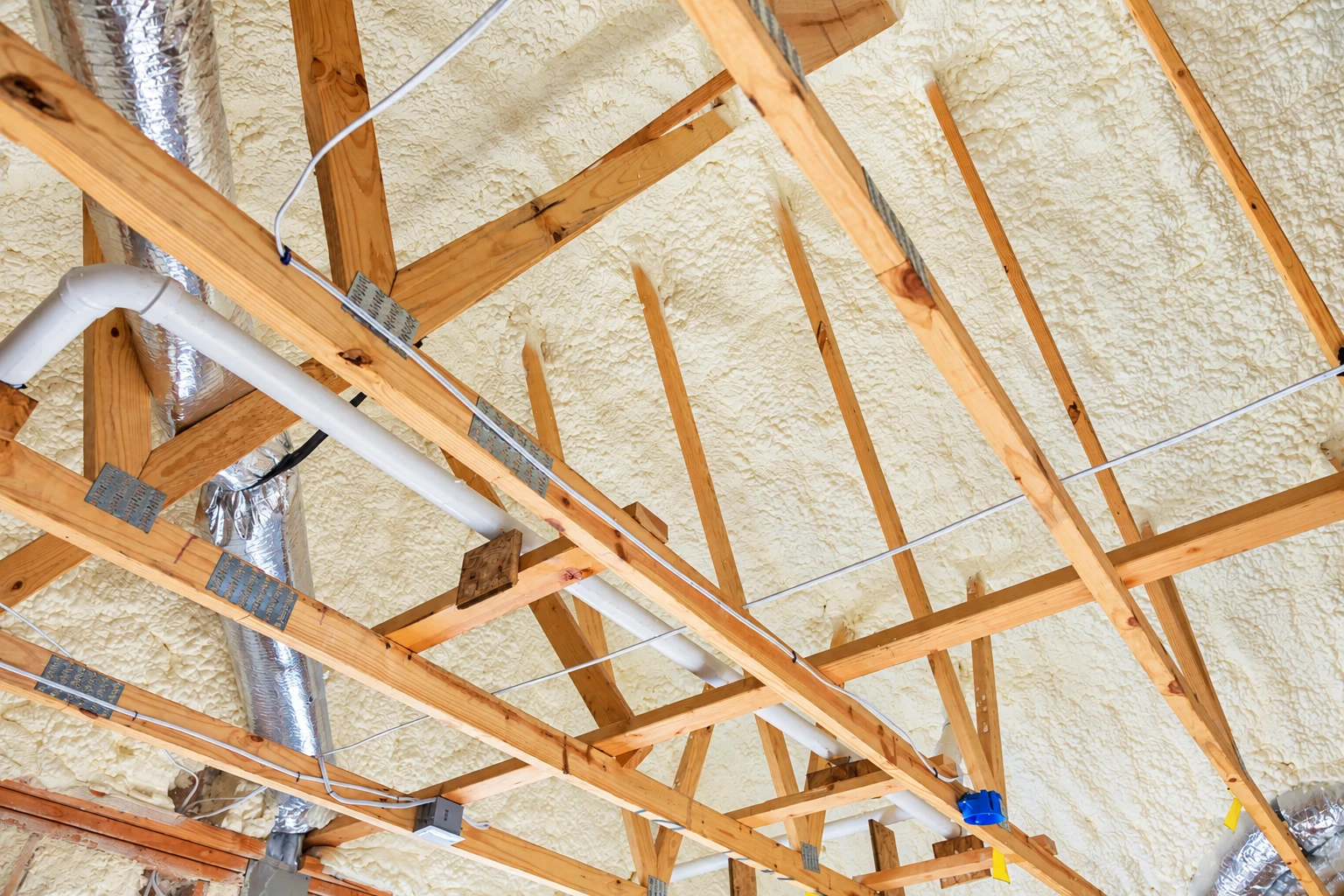 Wooden roof trusses cross a ceiling coated in pale spray foam, with ducts and wiring visible.