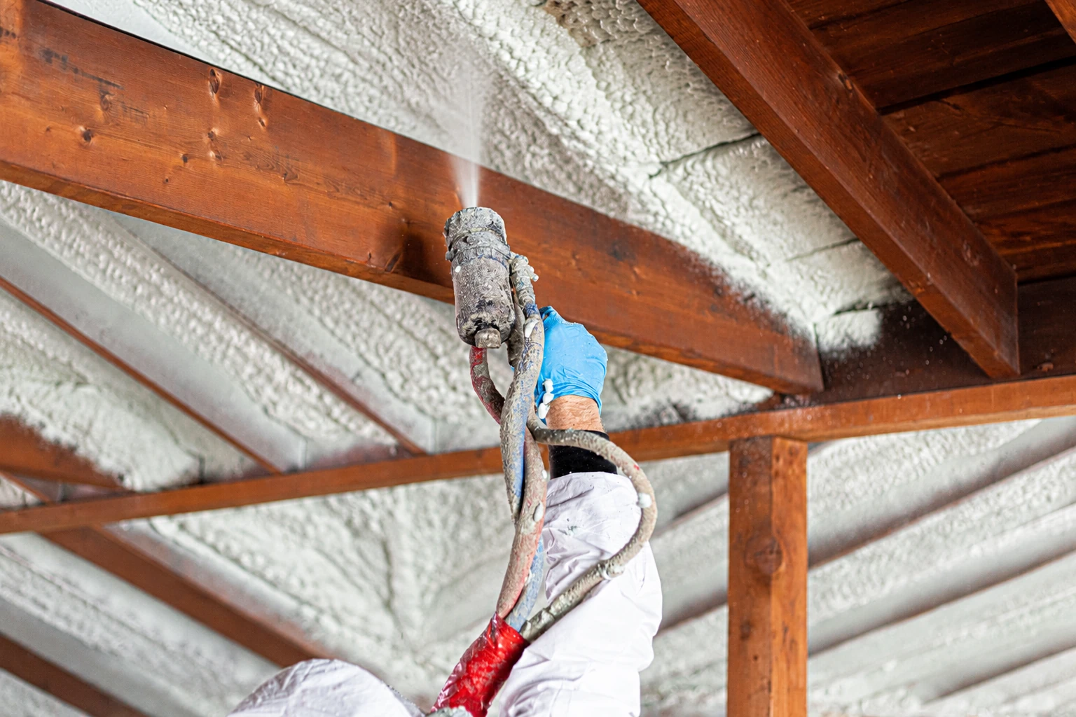 Worker sprays white foam insulation onto the underside of a wooden roof, holding a spray gun among exposed beams.