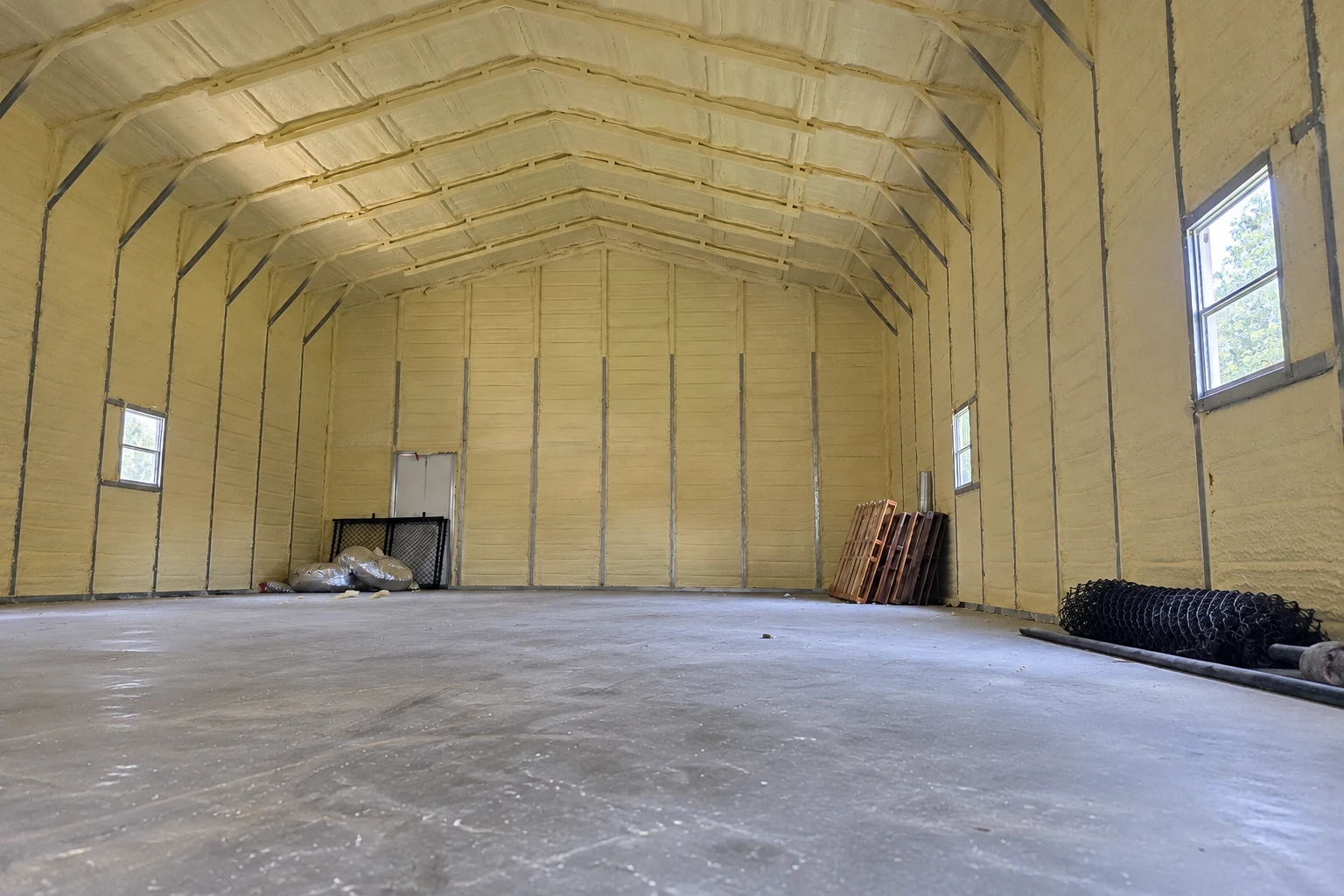 A large commercial building has pale spray foam covering its arched ceiling and walls, with windows and a bare concrete floor.