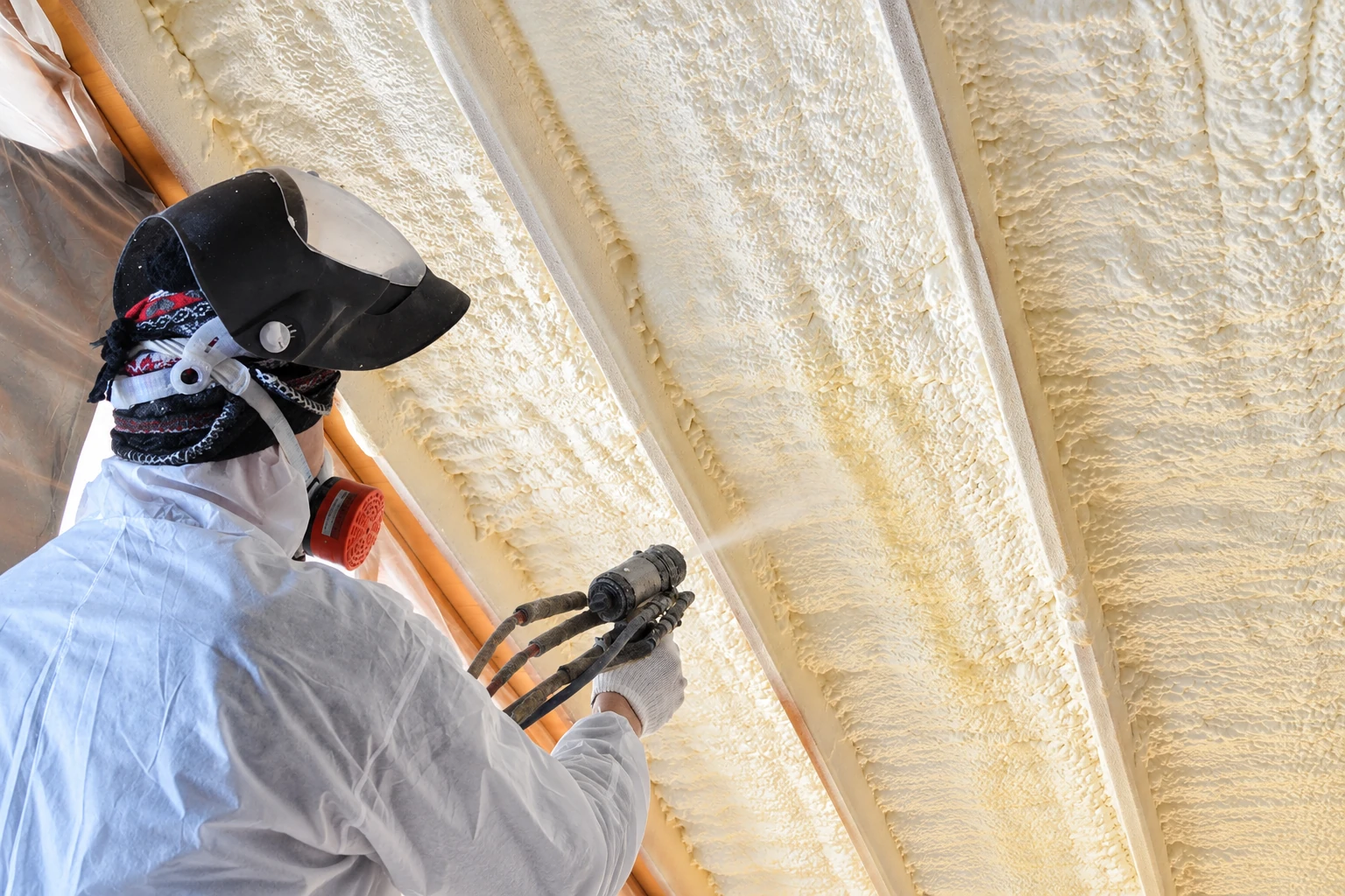 A worker in protective gear sprays foam insulation onto the underside of a sloped roof between rafters.