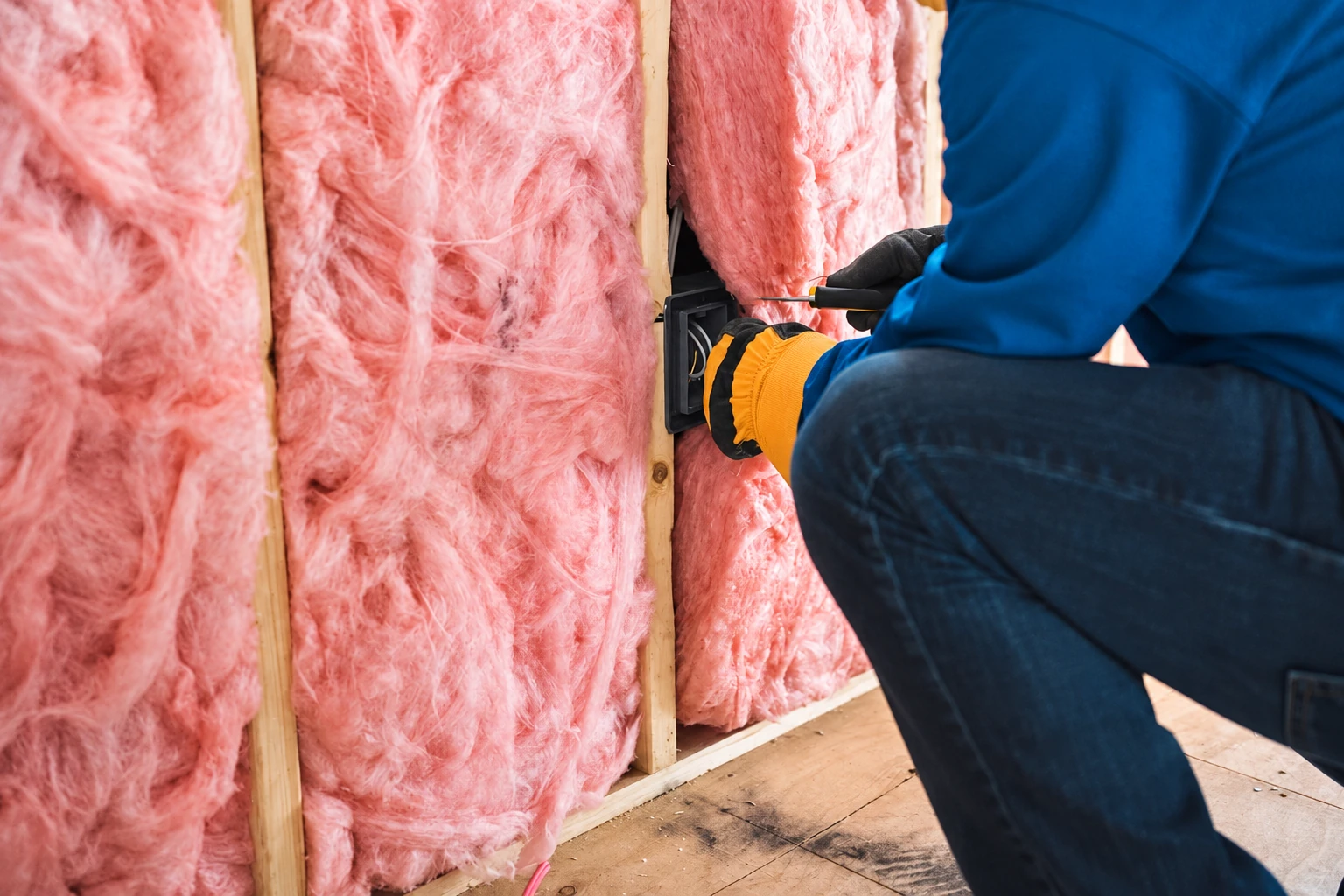 Worker fits pink fiberglass batts between wall studs beside an electrical outlet.
