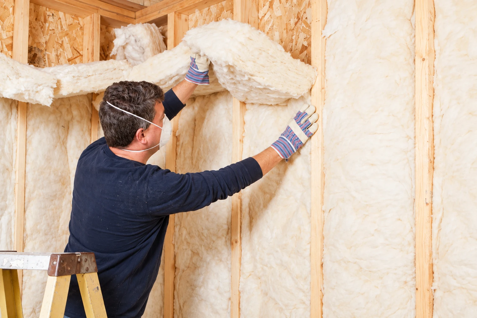A worker in a mask and gloves fits fluffy fiberglass batts between exposed wall studs.